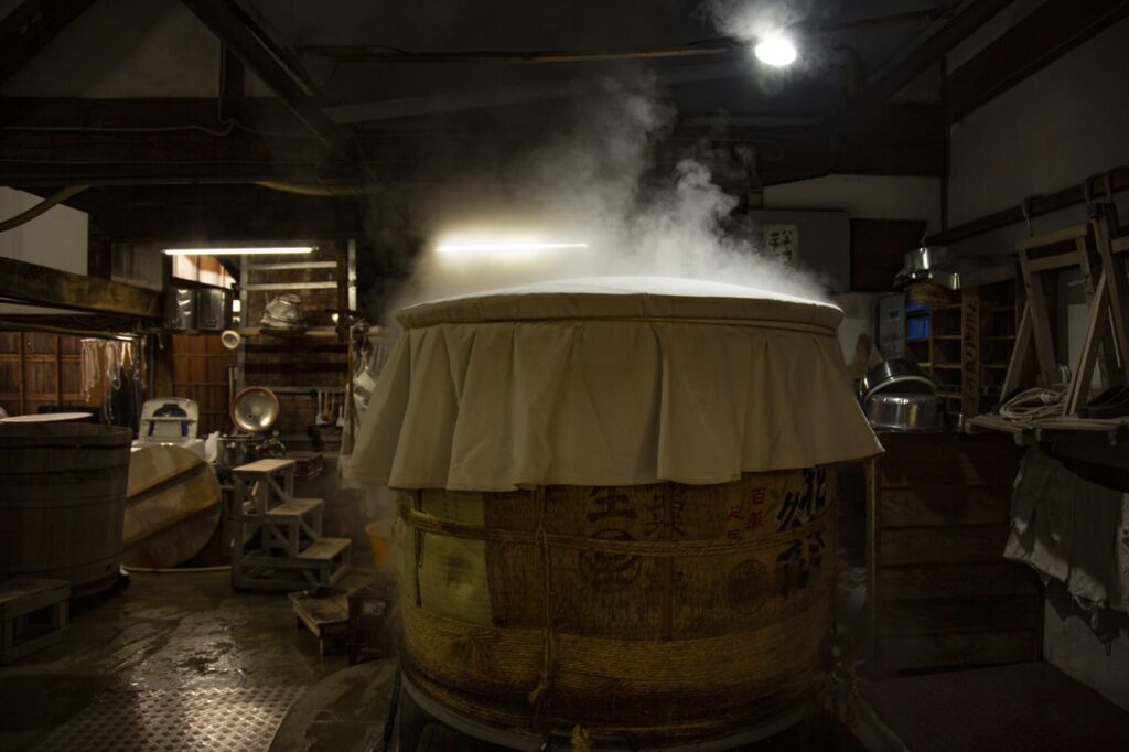 White steam billows from the koshiki (a large vat for steaming rice), causing the cloth cover to swell.